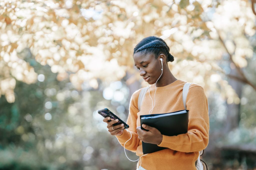 Young Black woman walking outside with headphones in, holding a folder and looking down at her phone